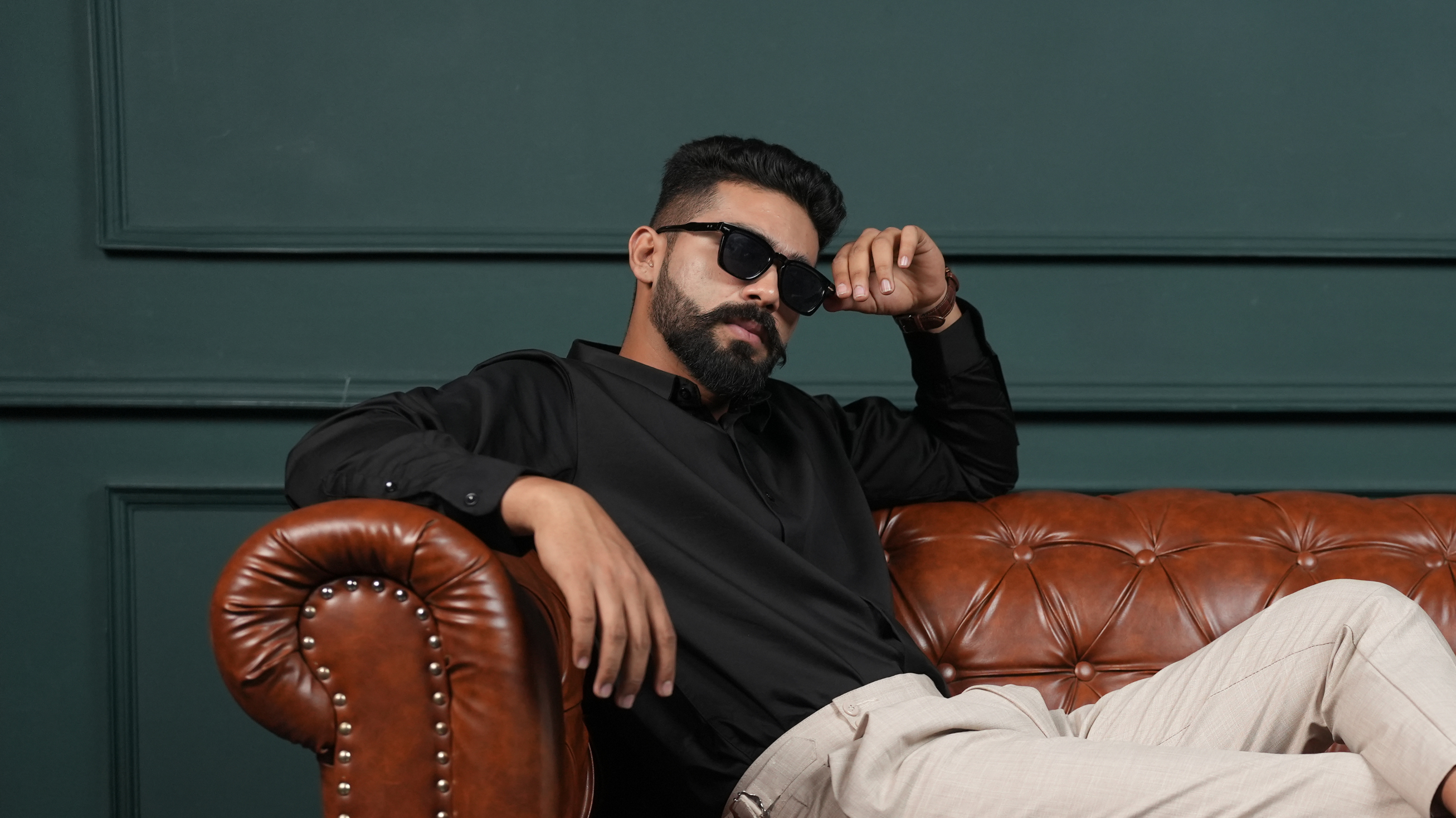 Man wearing hugrey classic shirt, sitting on a brown leather couch against a dark green wall.